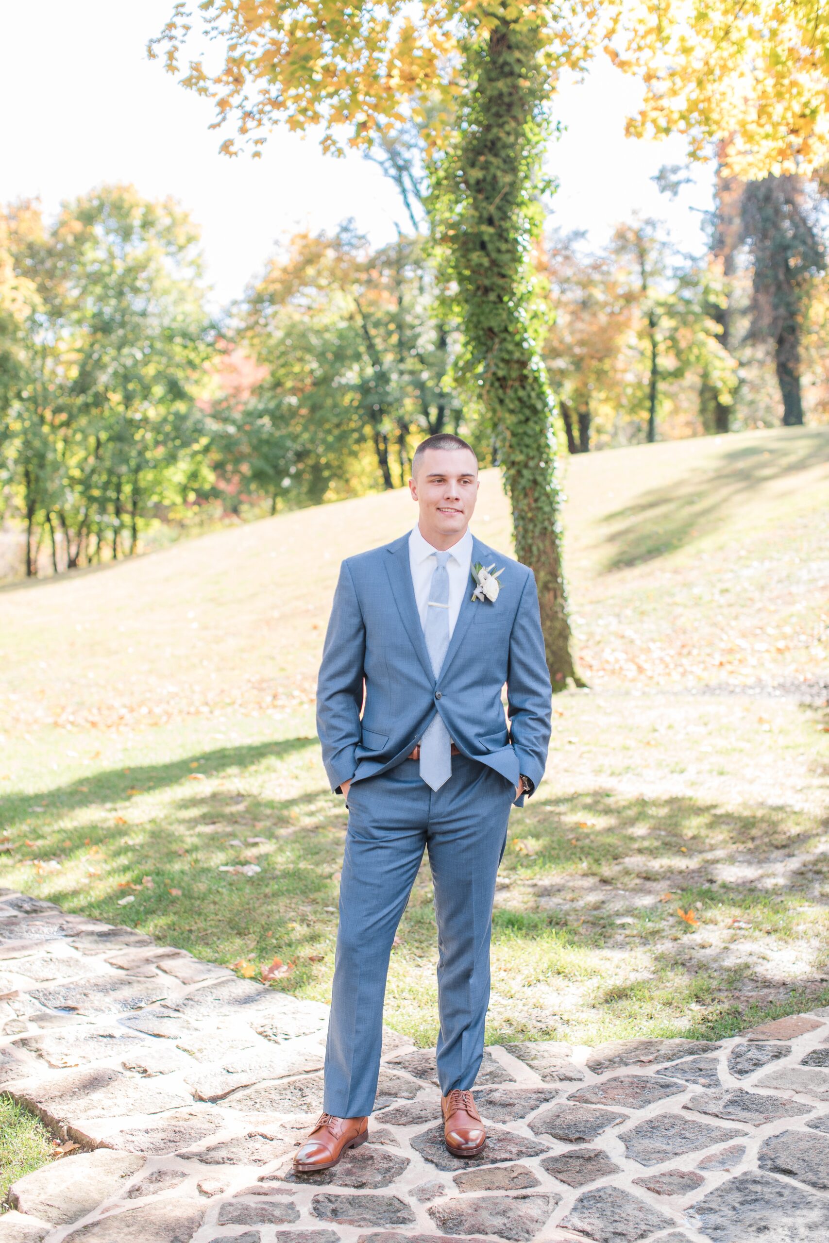 A groom waits on a stone sidewalk with hands in his blue pants pockets