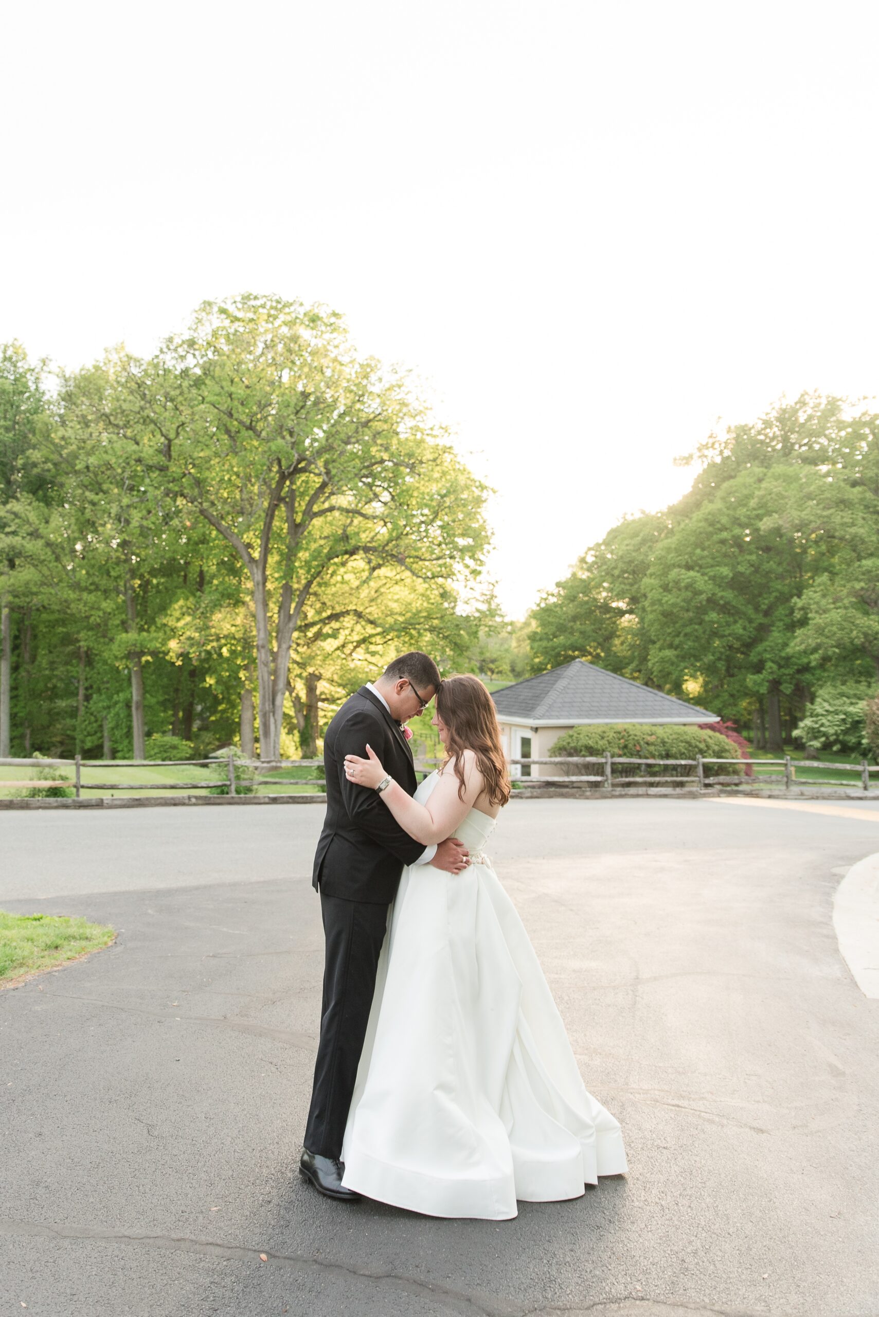 A bride and groom touch foreheads while standing in the driveway of the Manor Country Club Wedding venue at sunset