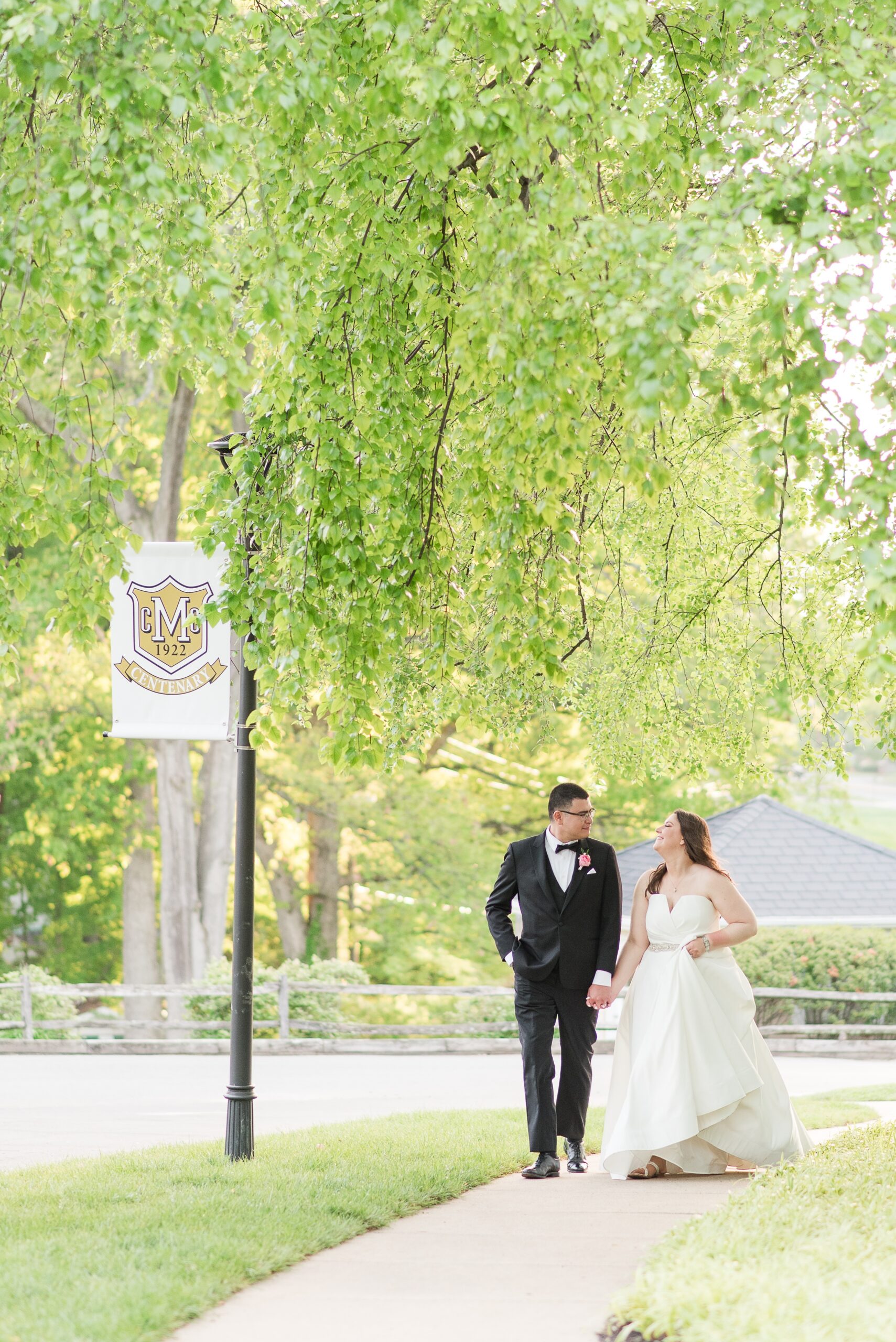 Newlyweds chat and laugh while walking under trees and holding hands at the Manor Country Club Wedding venue