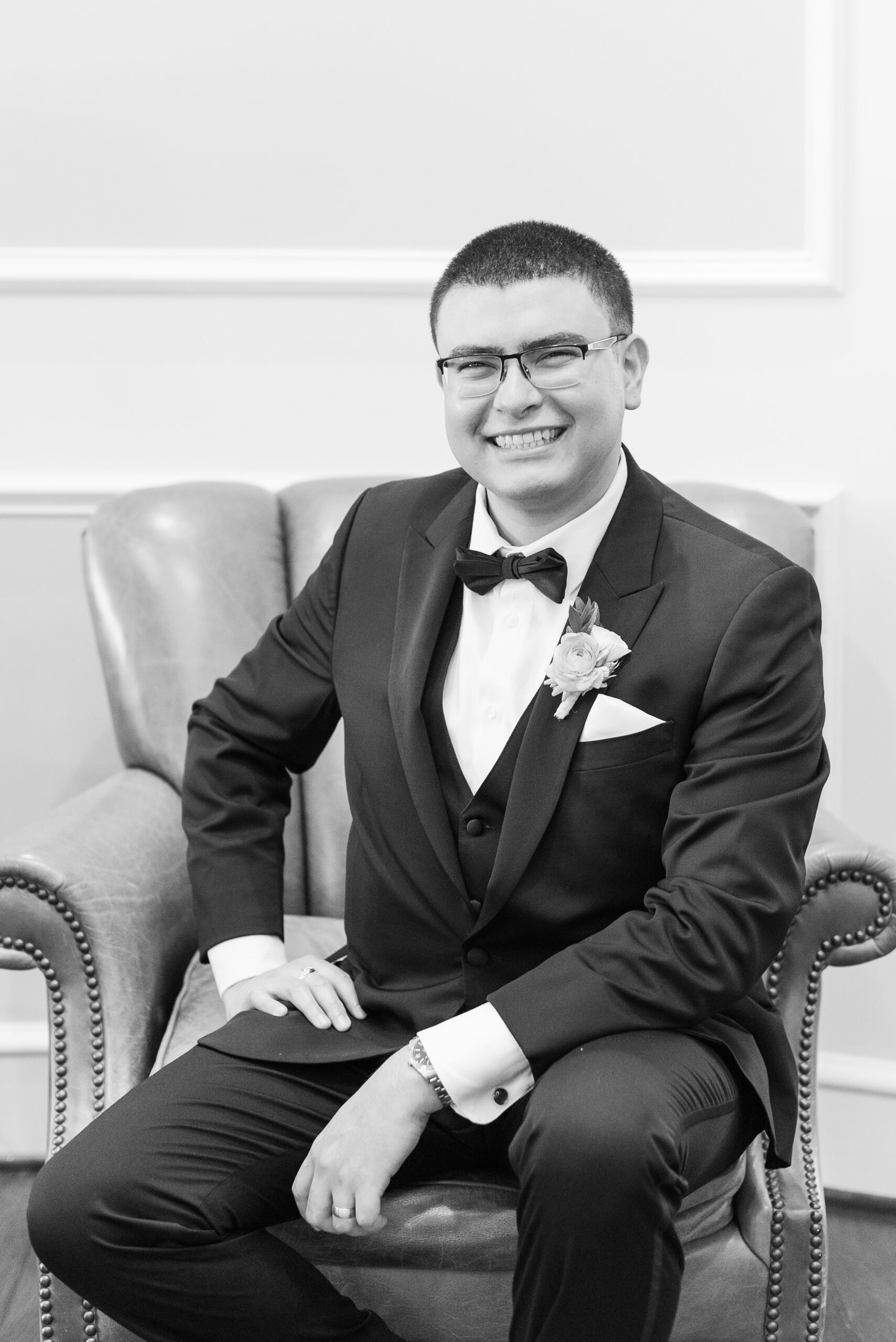 A groom sits in a leather chair smiling happily in his tuxedo