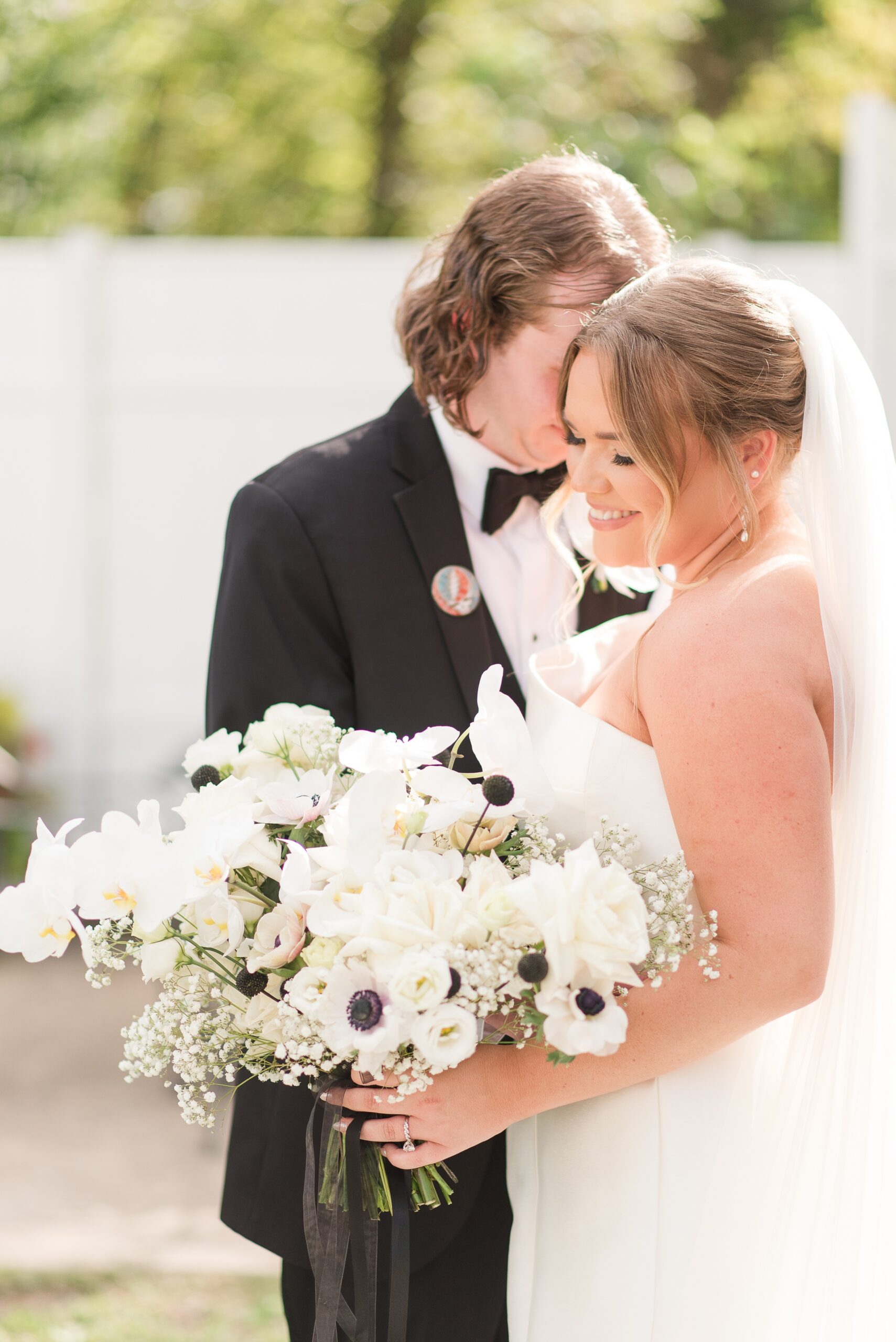 A bride smiles while standing with her groom and holding her white rose bouquet