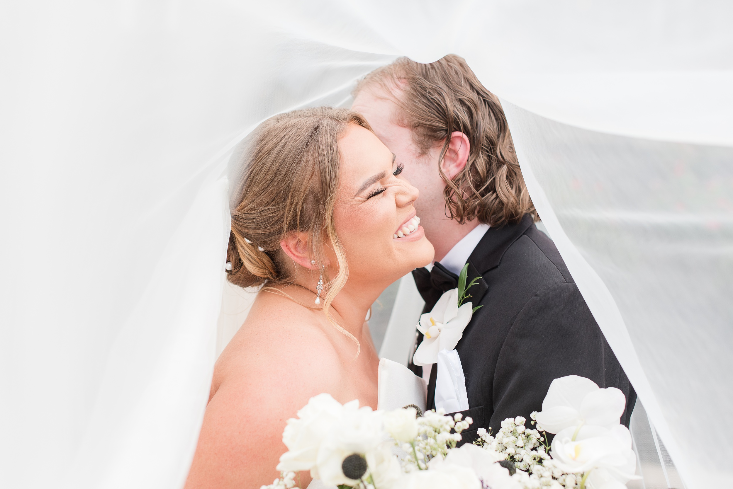 A bride giggles while her groom whispers in her ear under a veil