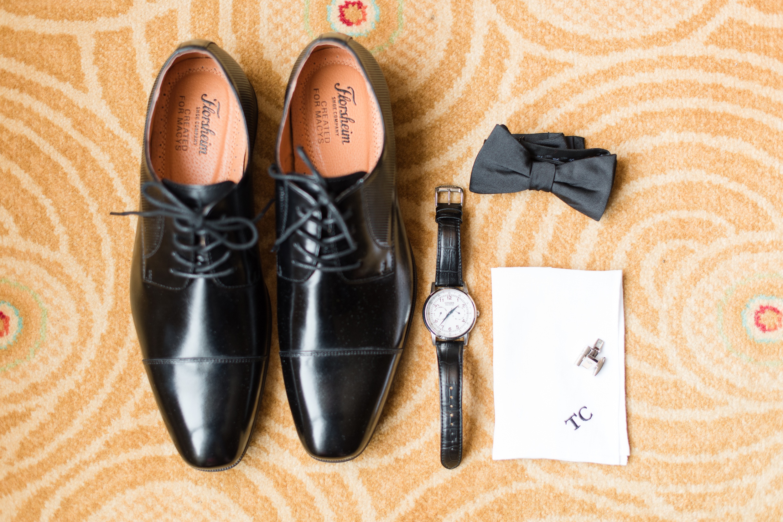 Details of a groom's black shoes, bowtie and watch sit on an orange rug together