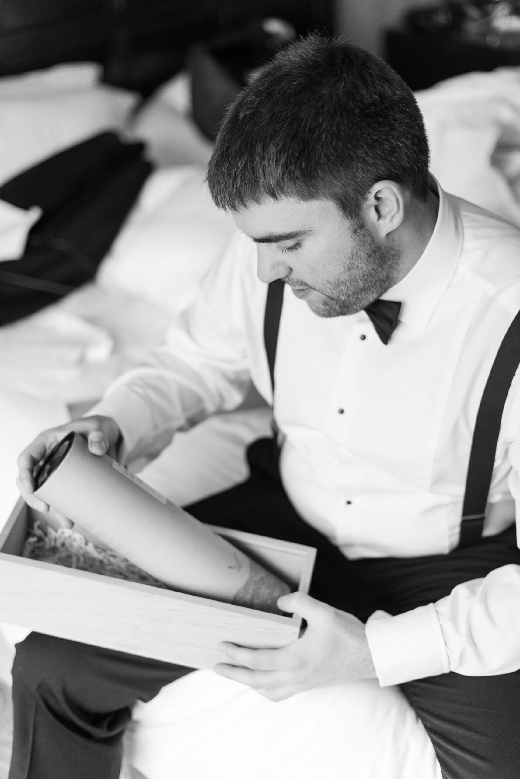 A groom opens a boxed gift while sitting on a bed without a jacket