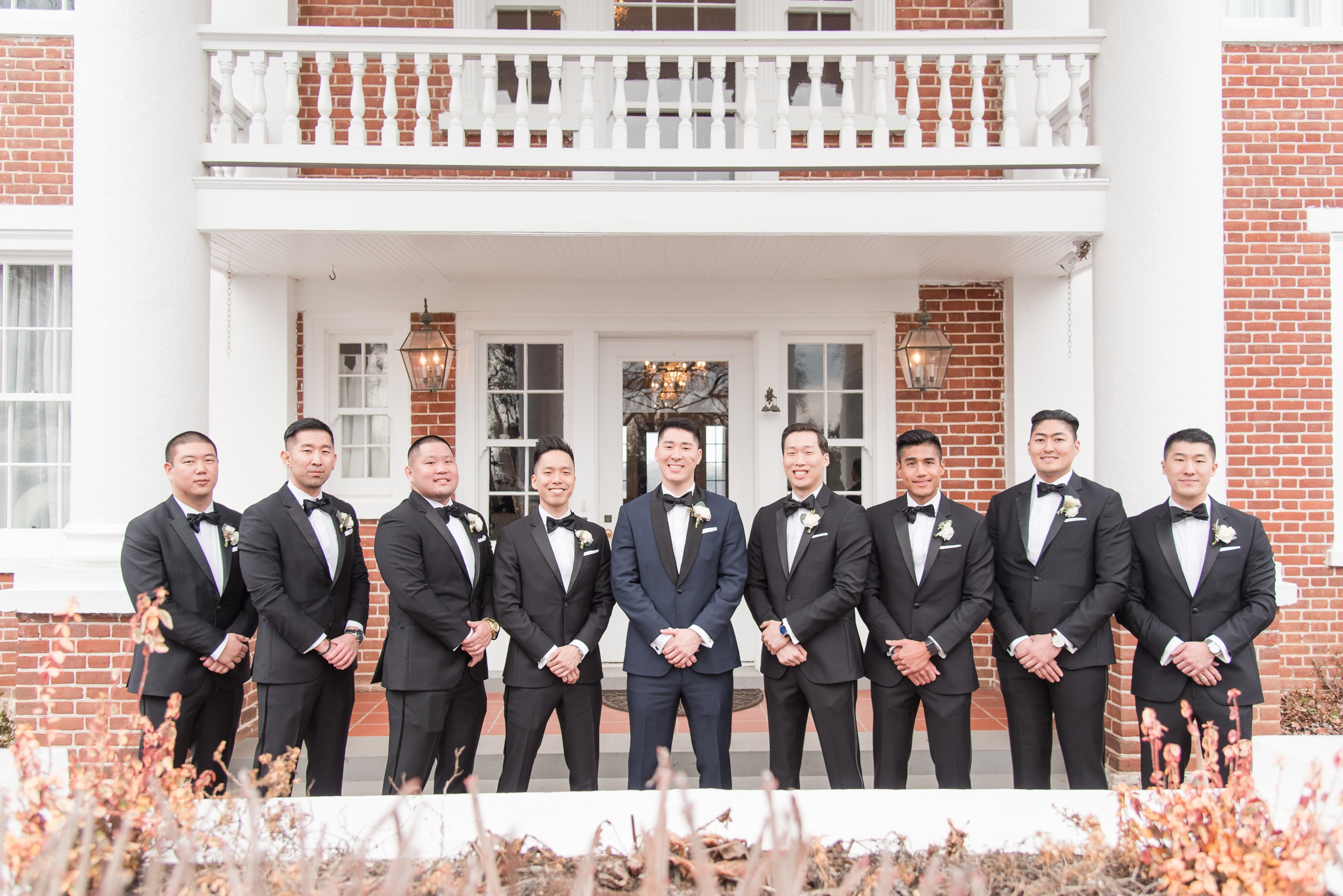 A groom stands under a balcony with his large party of groomsmen with hands crossed