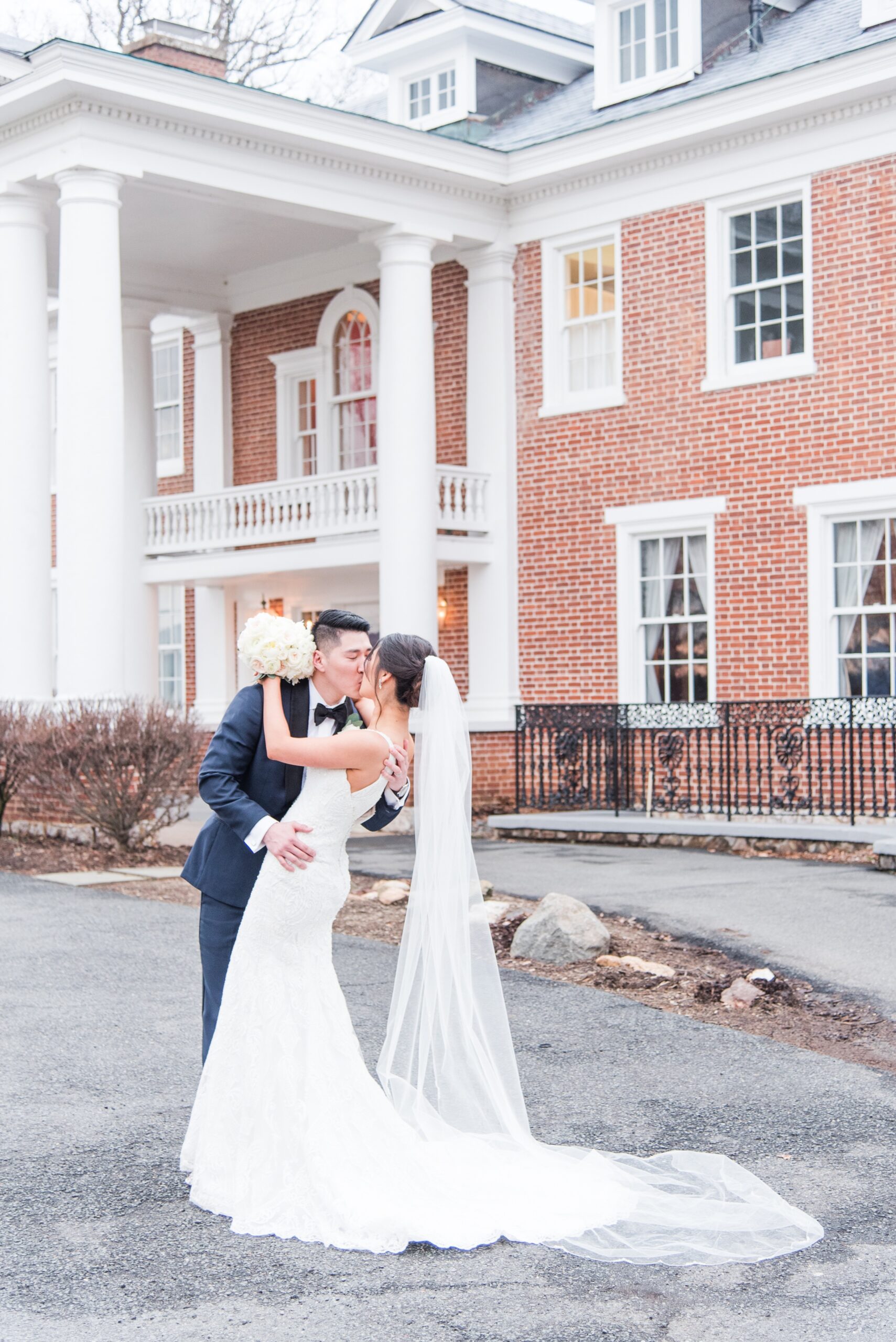 A groom in a blue suit dips his bride in the driveway of their wedding venue