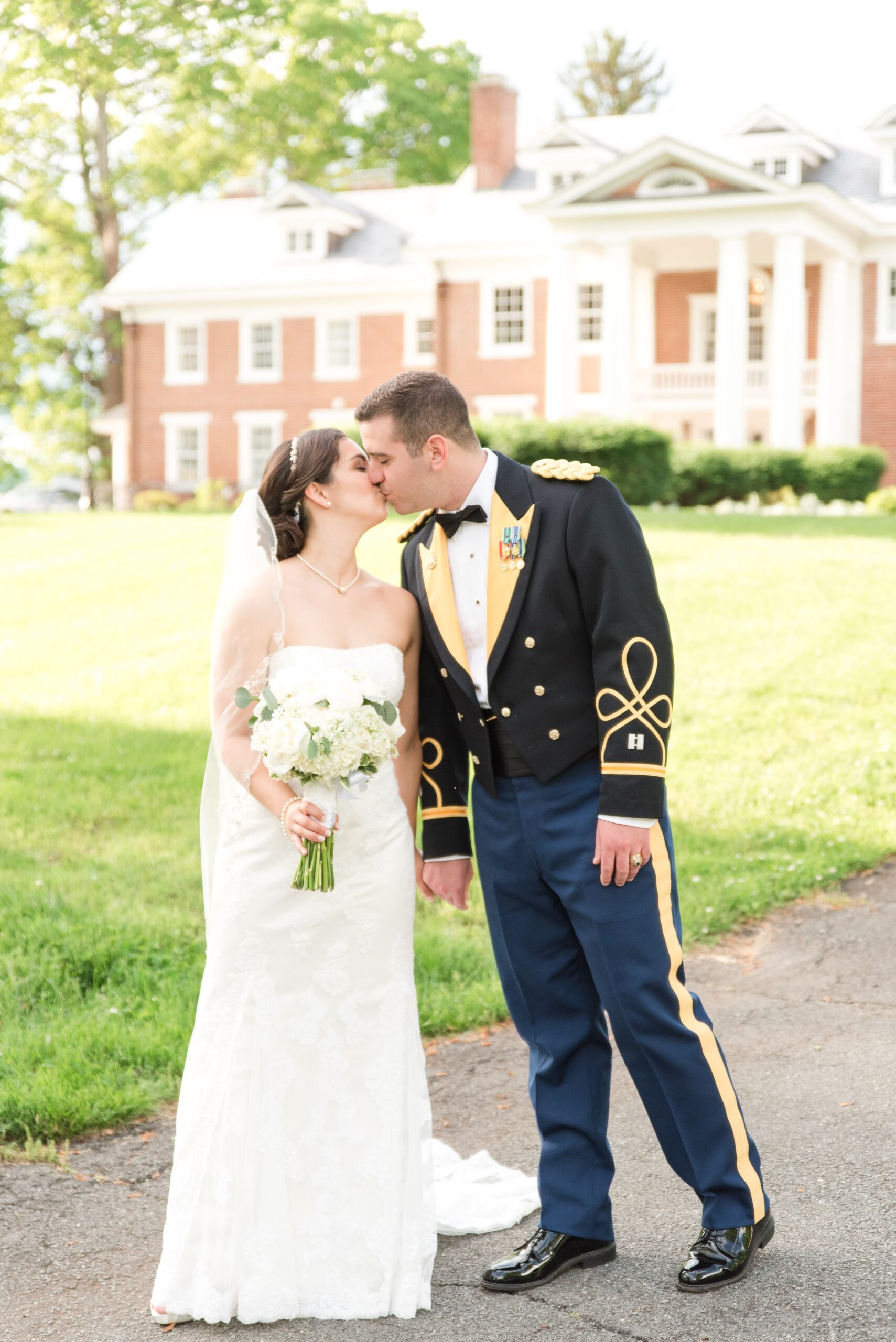 A groom in a military uniform kisses his bride in the lawn at their wedding