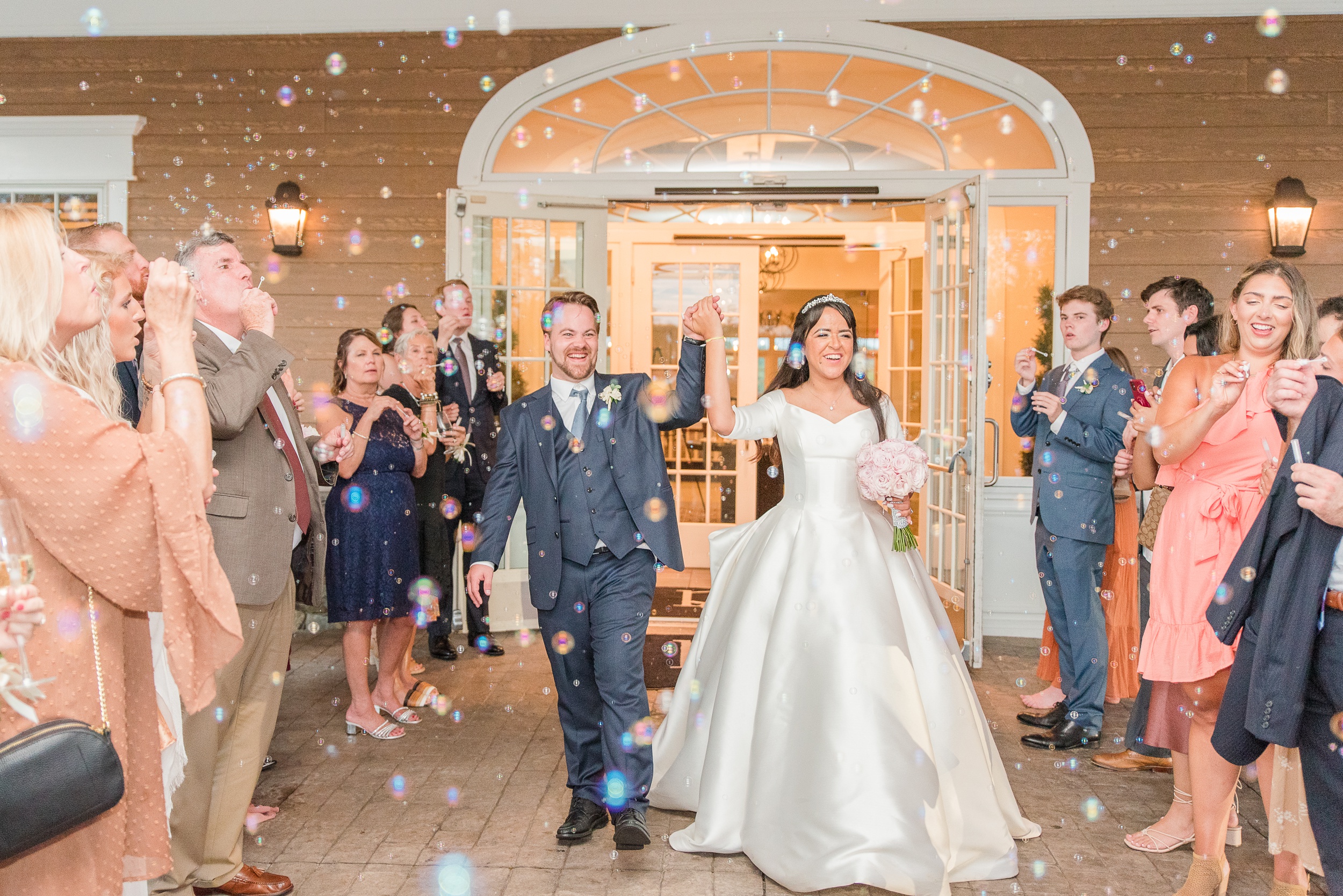 Newlyweds raise their hands while exiting their wedding reception under bubbles at the River Creek Club Wedding venue