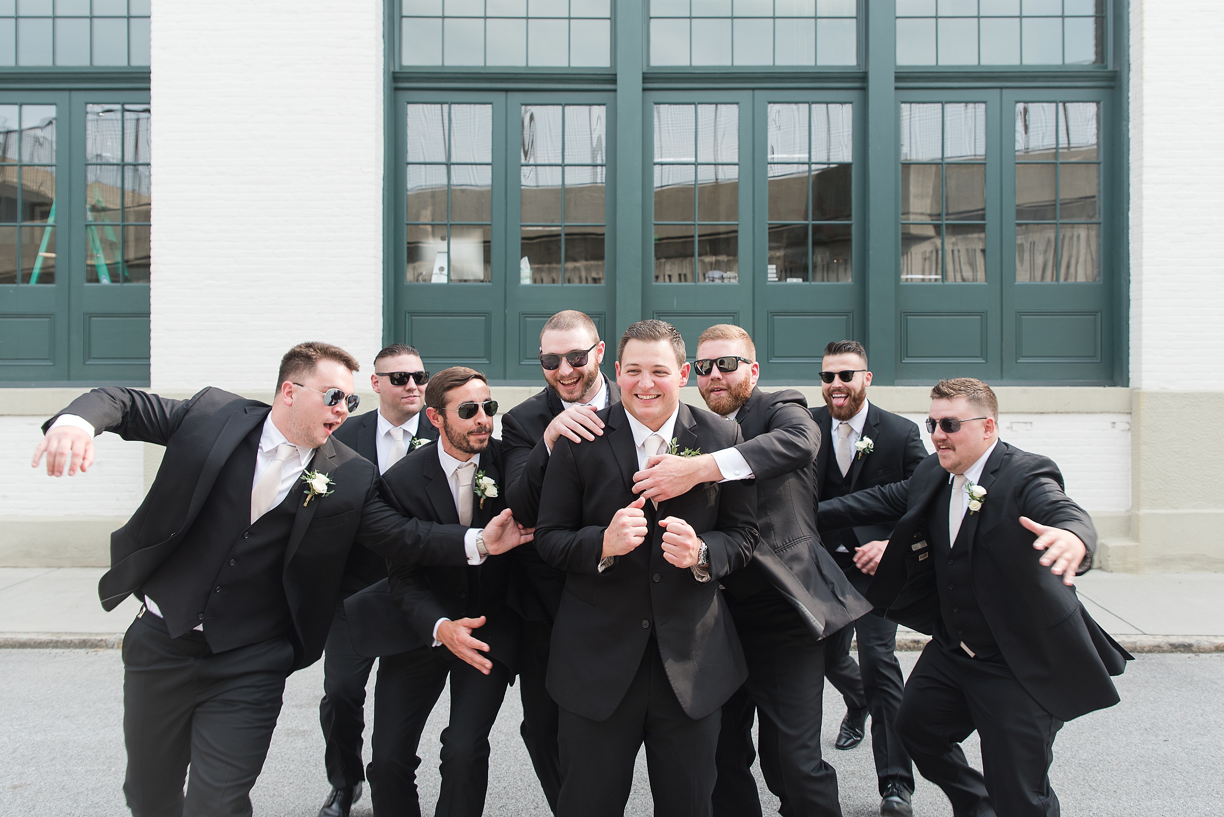 A groom is hugged and attacked by his groomsmen in all black suits and sunglasses