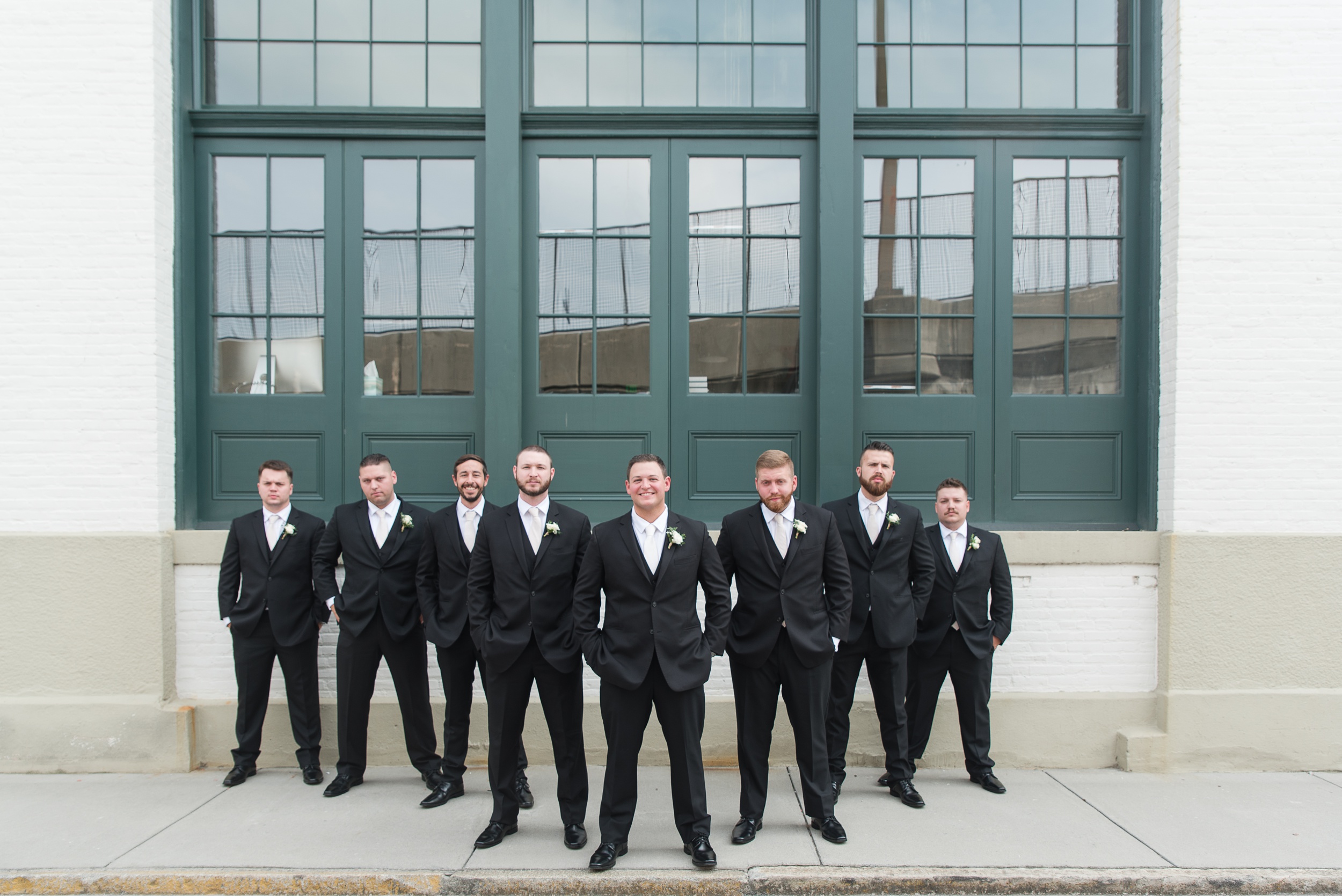 A groom stands with hands in his pockets with his groomsman in black suits with white ties