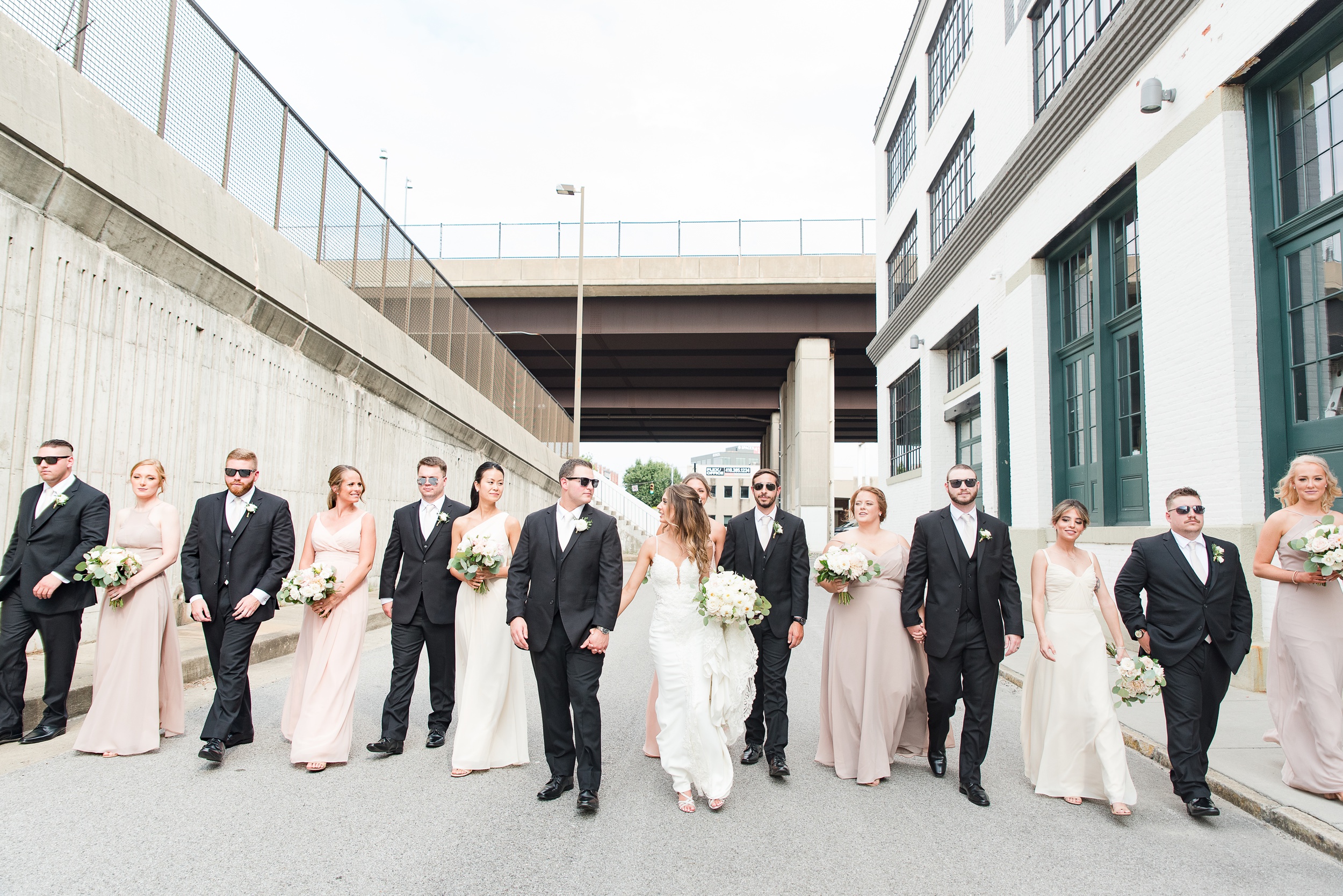 A bride and groom hold hands while walking through an alley with their wedding party at The Winslow Baltimore