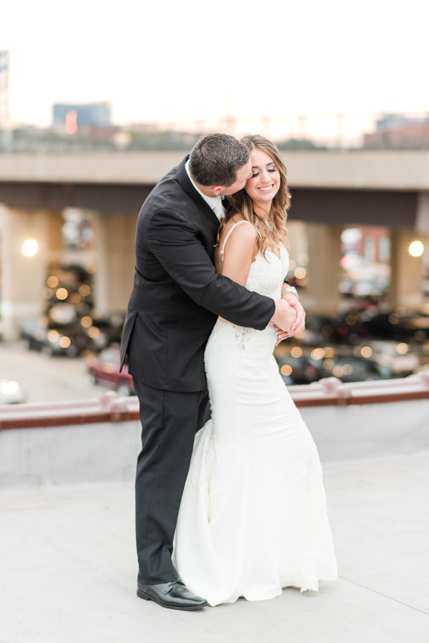 A groom hugs and kisses his bride while standing on a rooftop at The Winslow Baltimore