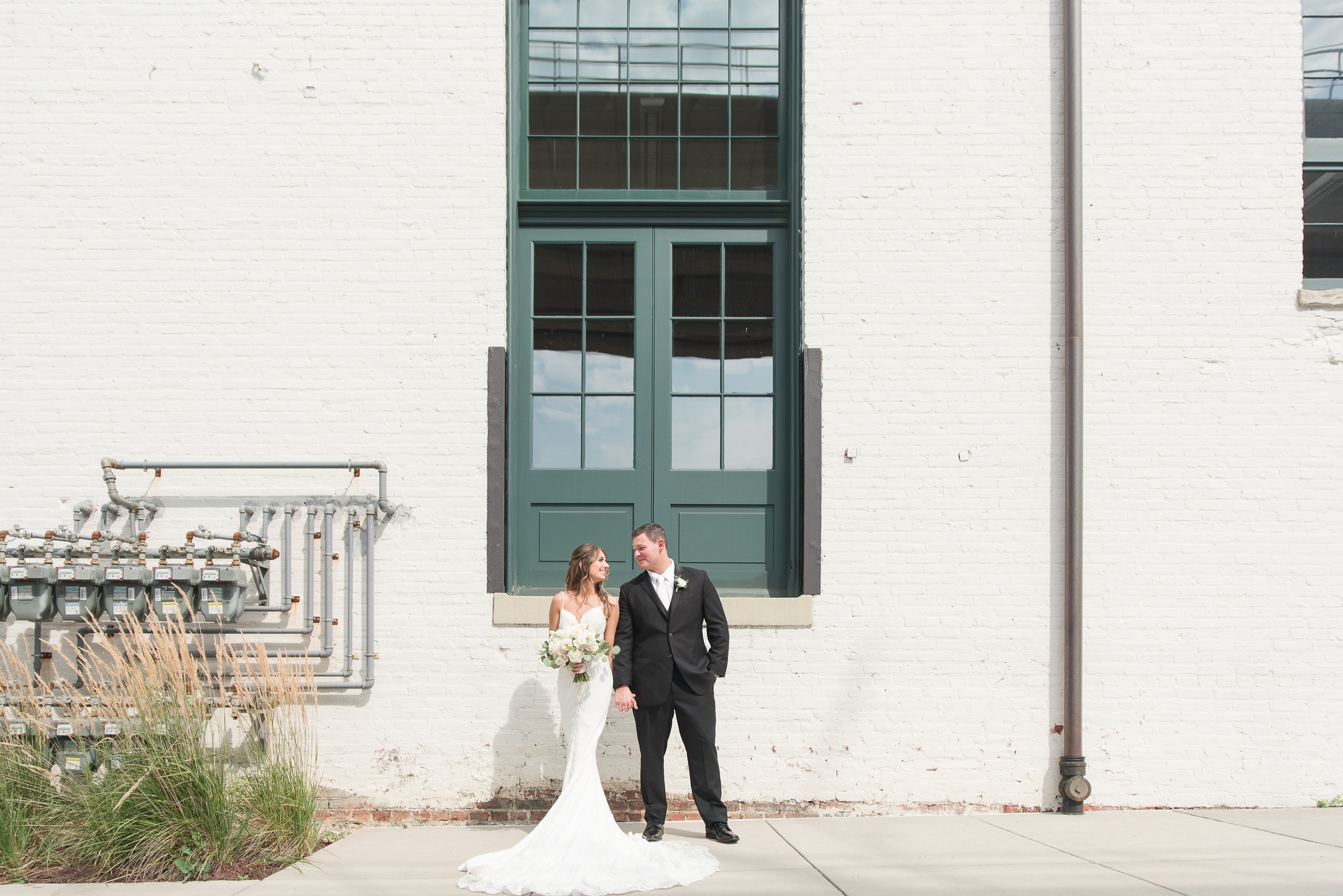 Newlyweds hold hands while walking in an alley by a white washed industrial building at The Winslow Baltimore