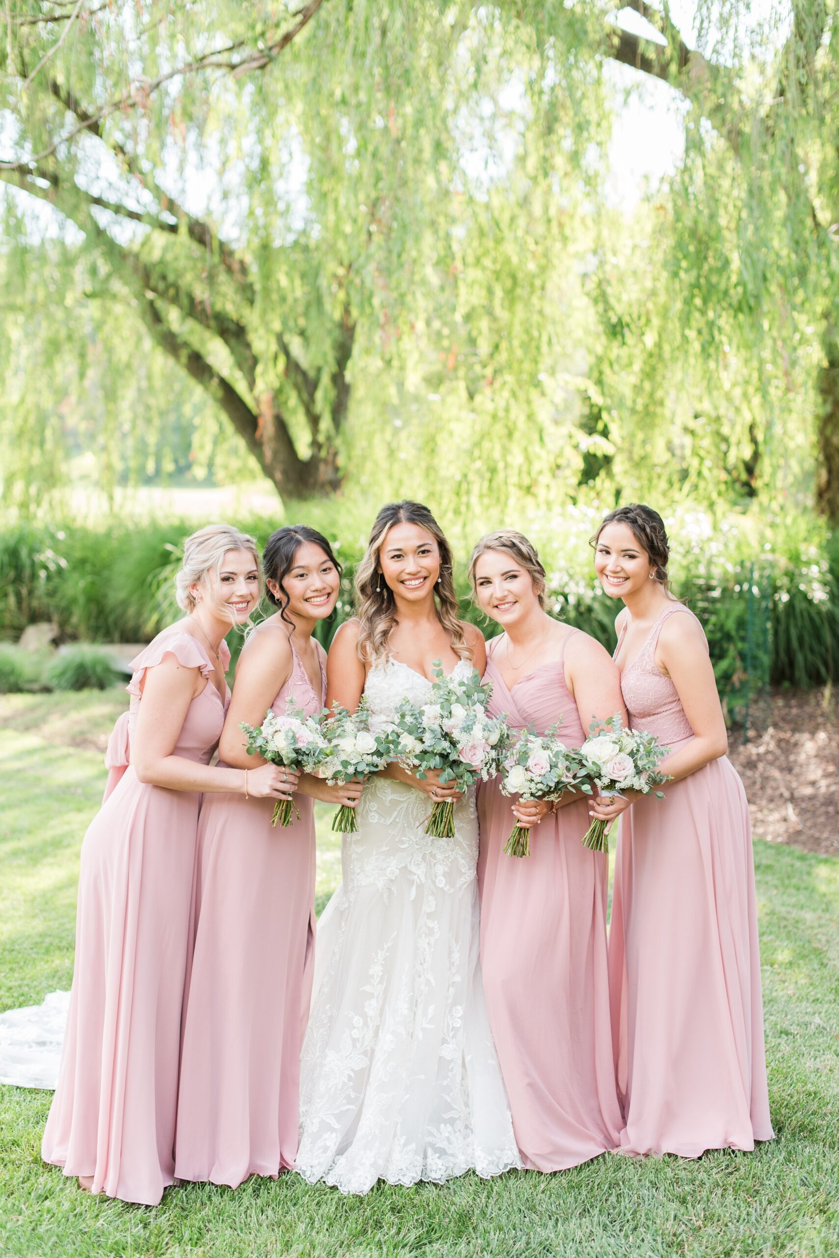 A bride and her bridesmaids in pink dresses stand together smiling and holding their bouquets under a willow tree in a garden