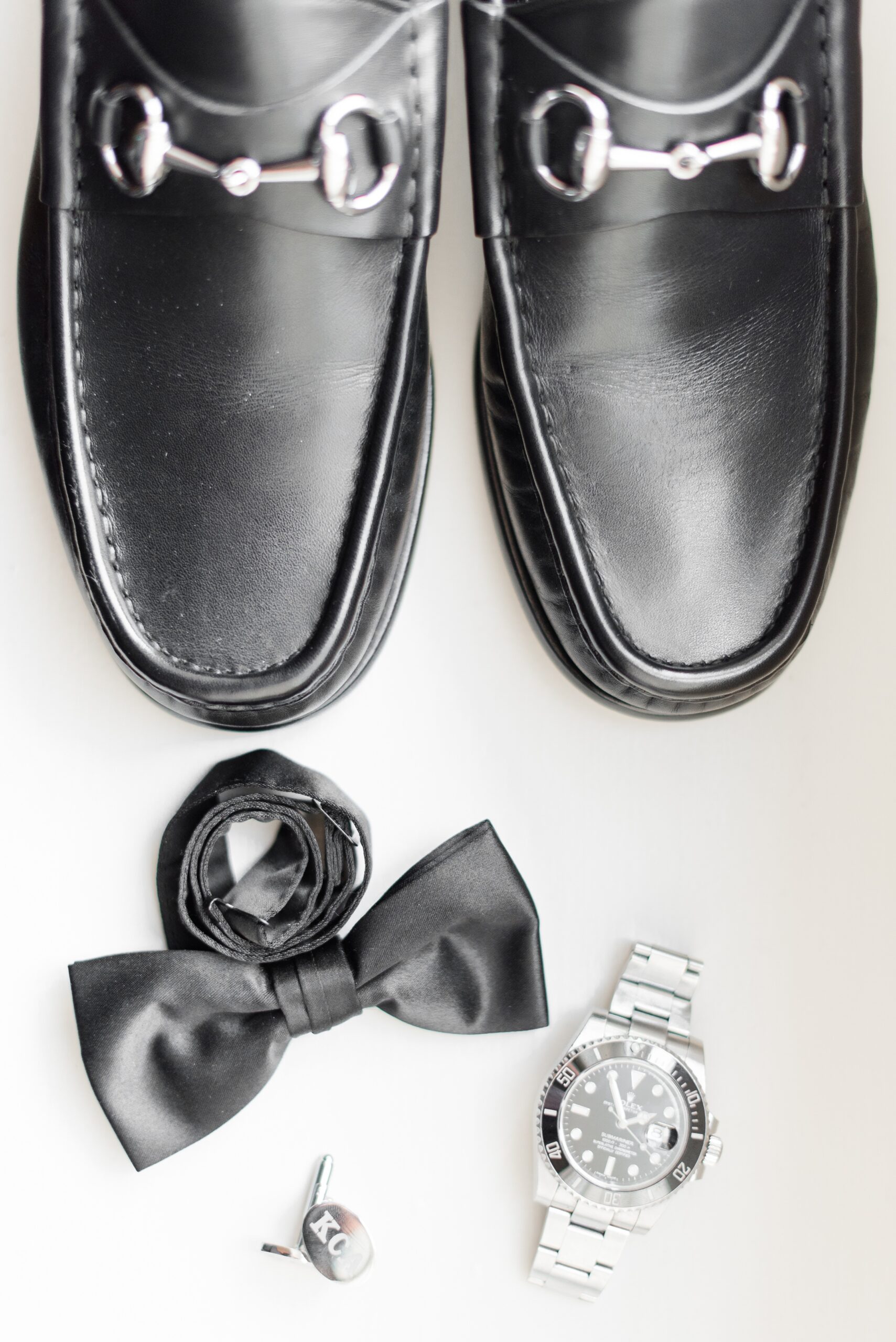 Details from the groom of black shoes and bowtie on a table with a watch and cufflinks