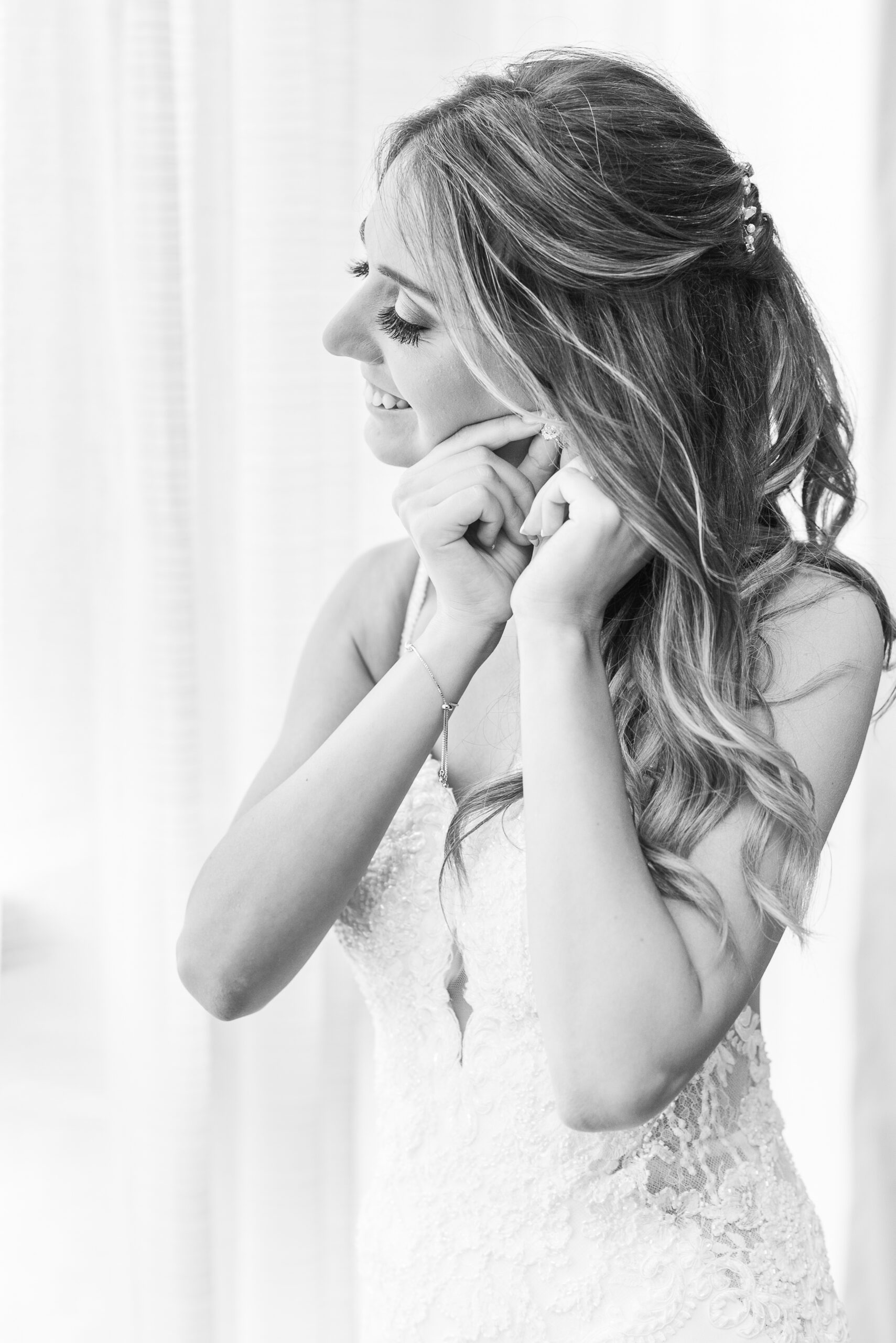 A bride in a lace dress smiles while putting on her diamond earrings at her Vandiver Inn Wedding