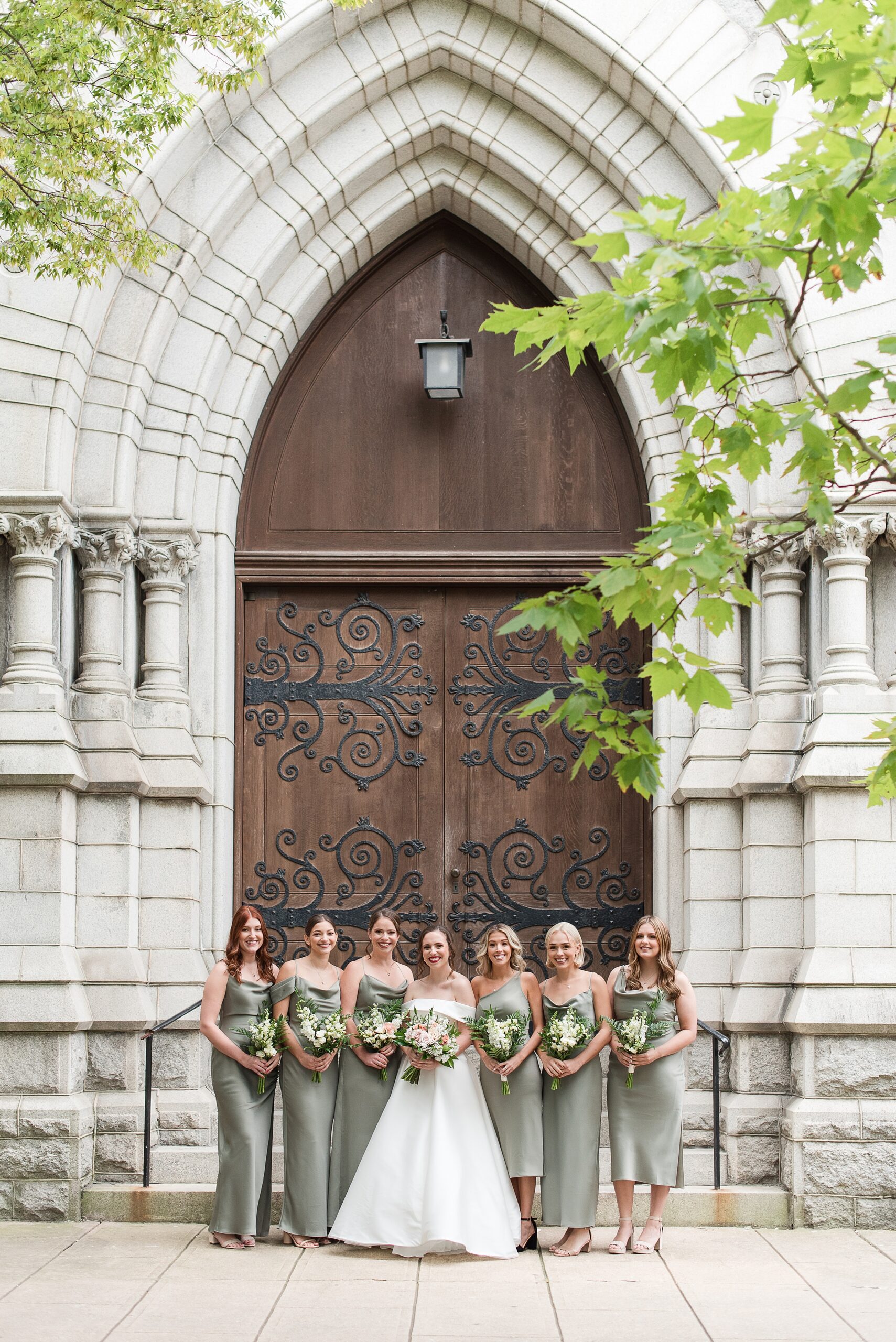 6 bridesmaids and a bride stand holding bouquets in front of large arched wooden doors to an ornate building