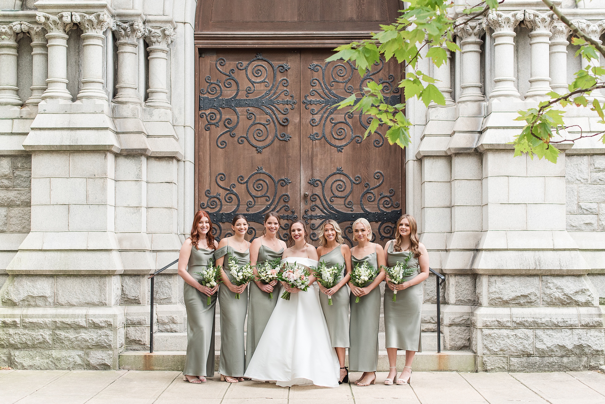 A bride smiles with her bridesmaids in green dresses while standing outside an ornate entrance to one of the incredible Wedding Venues Columbia, MD