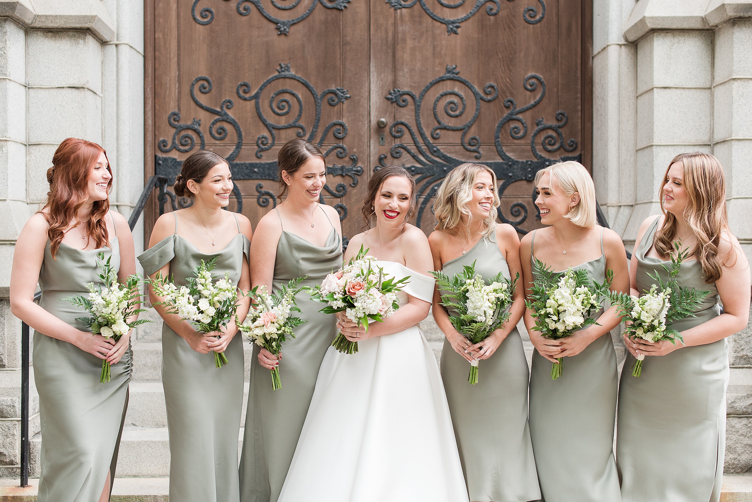 A bride stands with her bridesmaids in green dresses in front of a large wooden door entrance to Wedding Venues Columbia, MD