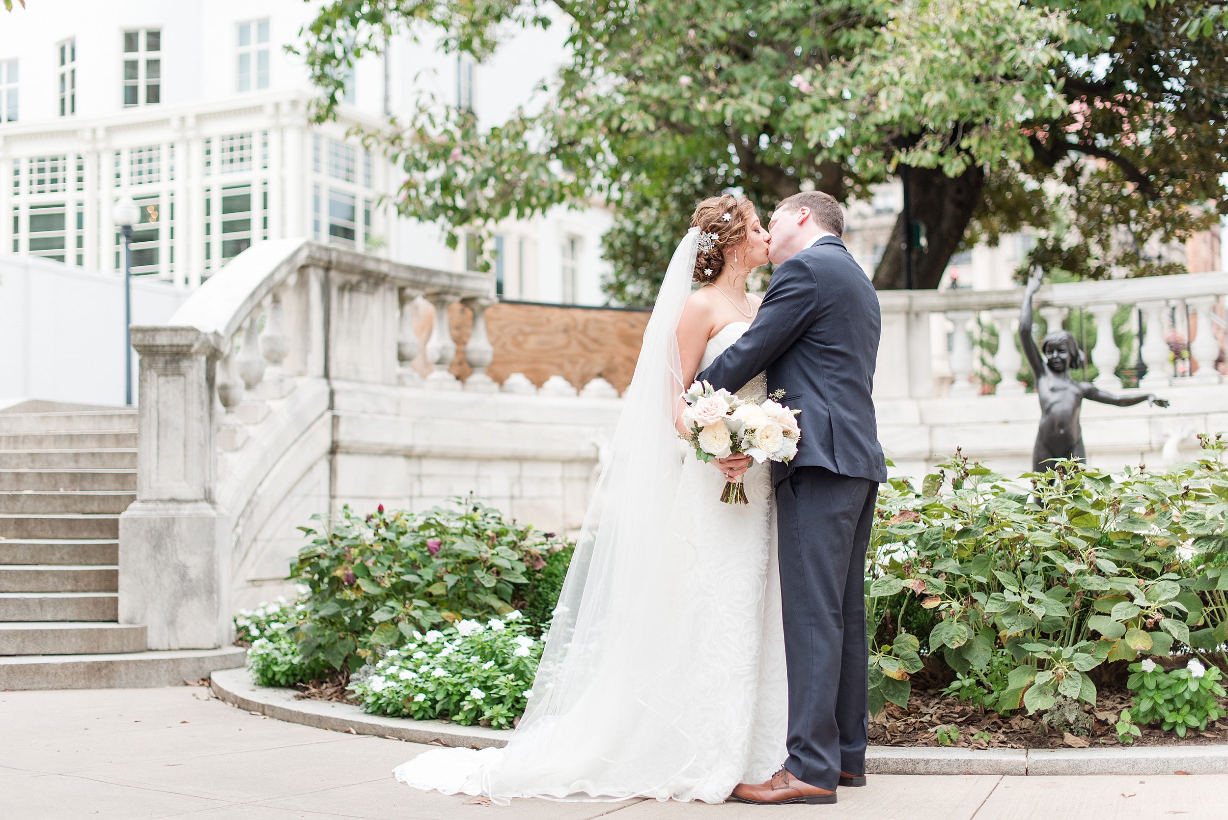 Newlyweds kiss in an ornate garden while holding a white rose bouquet at one of the Wedding Venues In Maryland