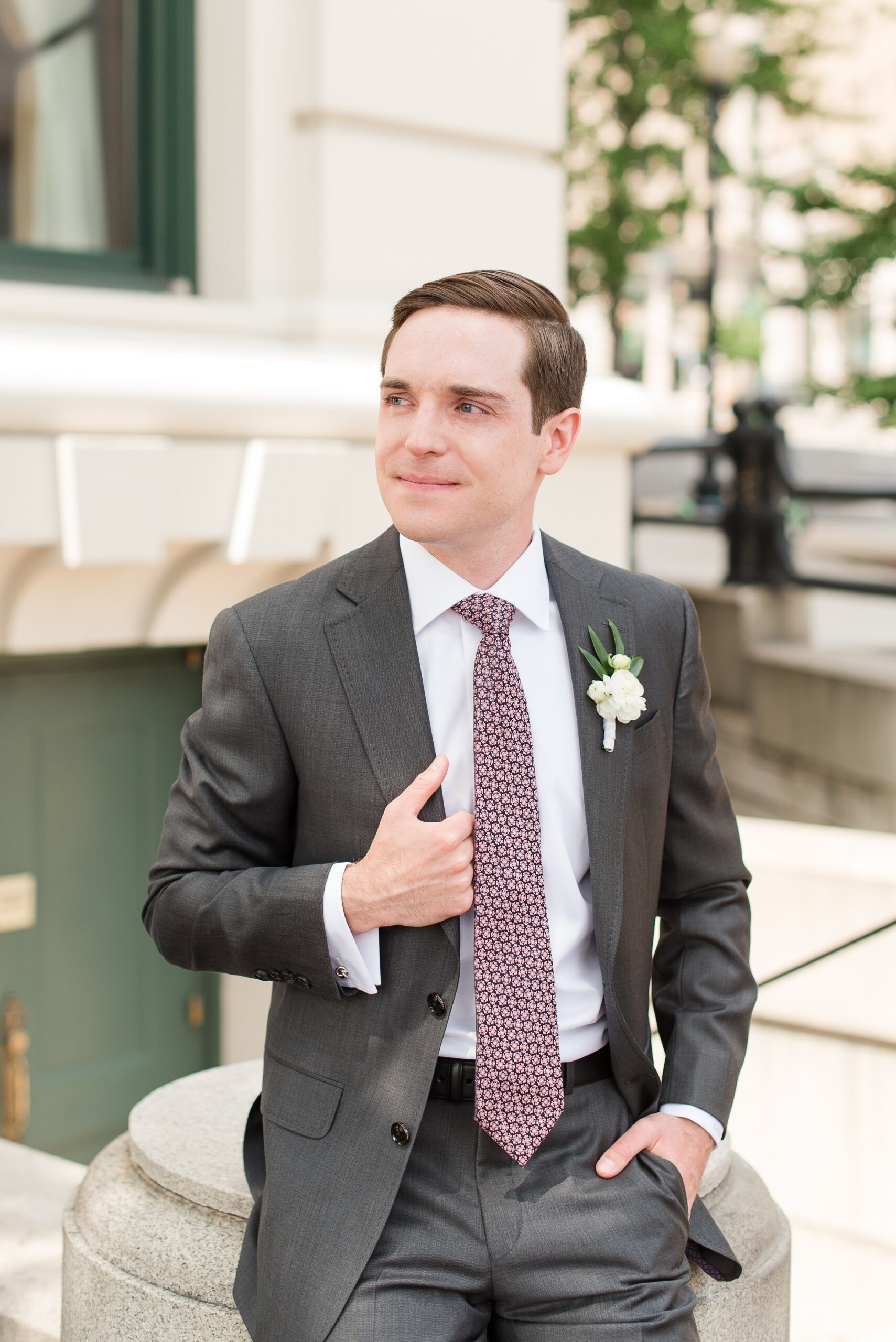 A groom smiles and holds his lapel while leaning on a concrete stair railing