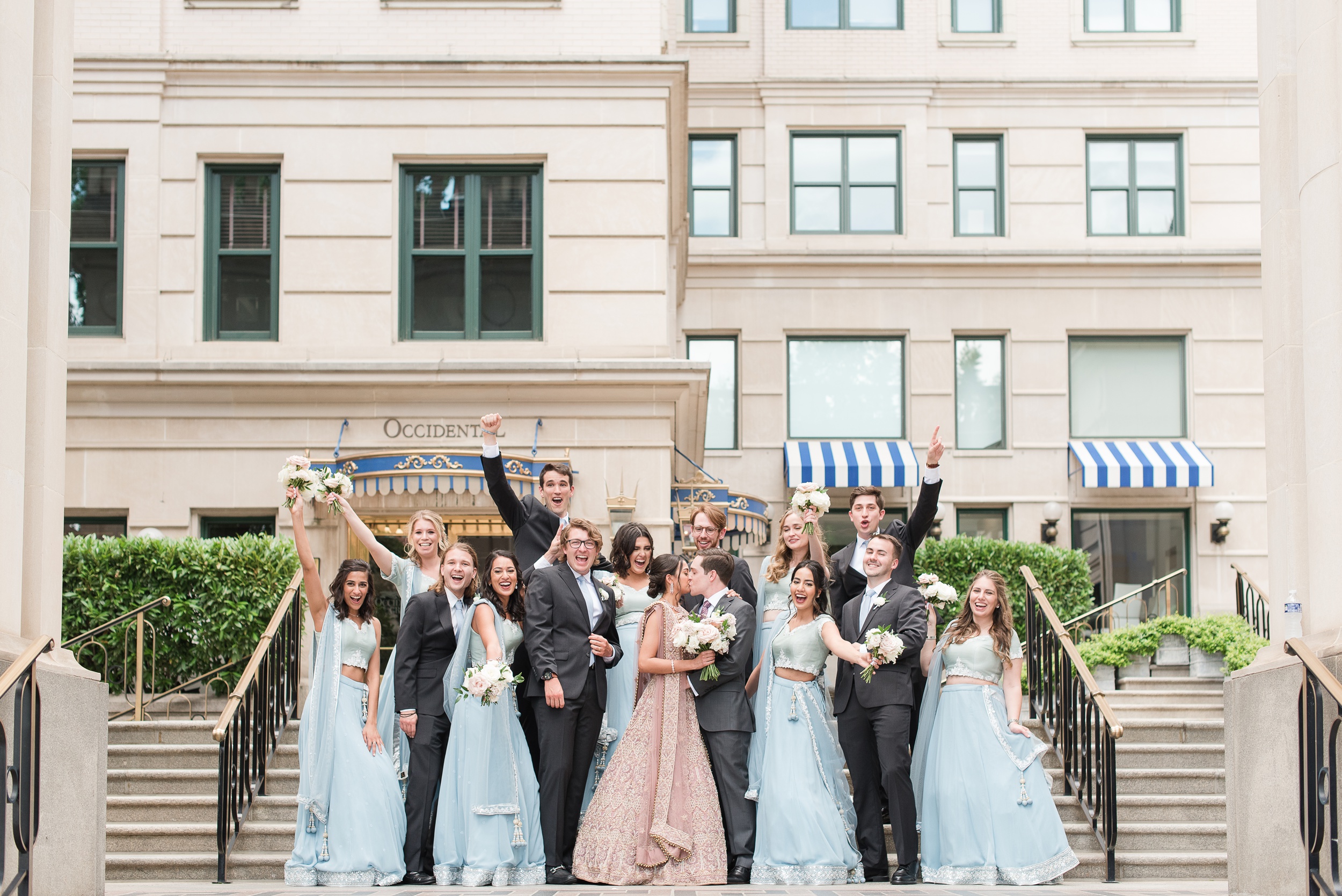 Newlyweds kiss while standing on stairs with their celebrating wedding party at their DAR Wedding