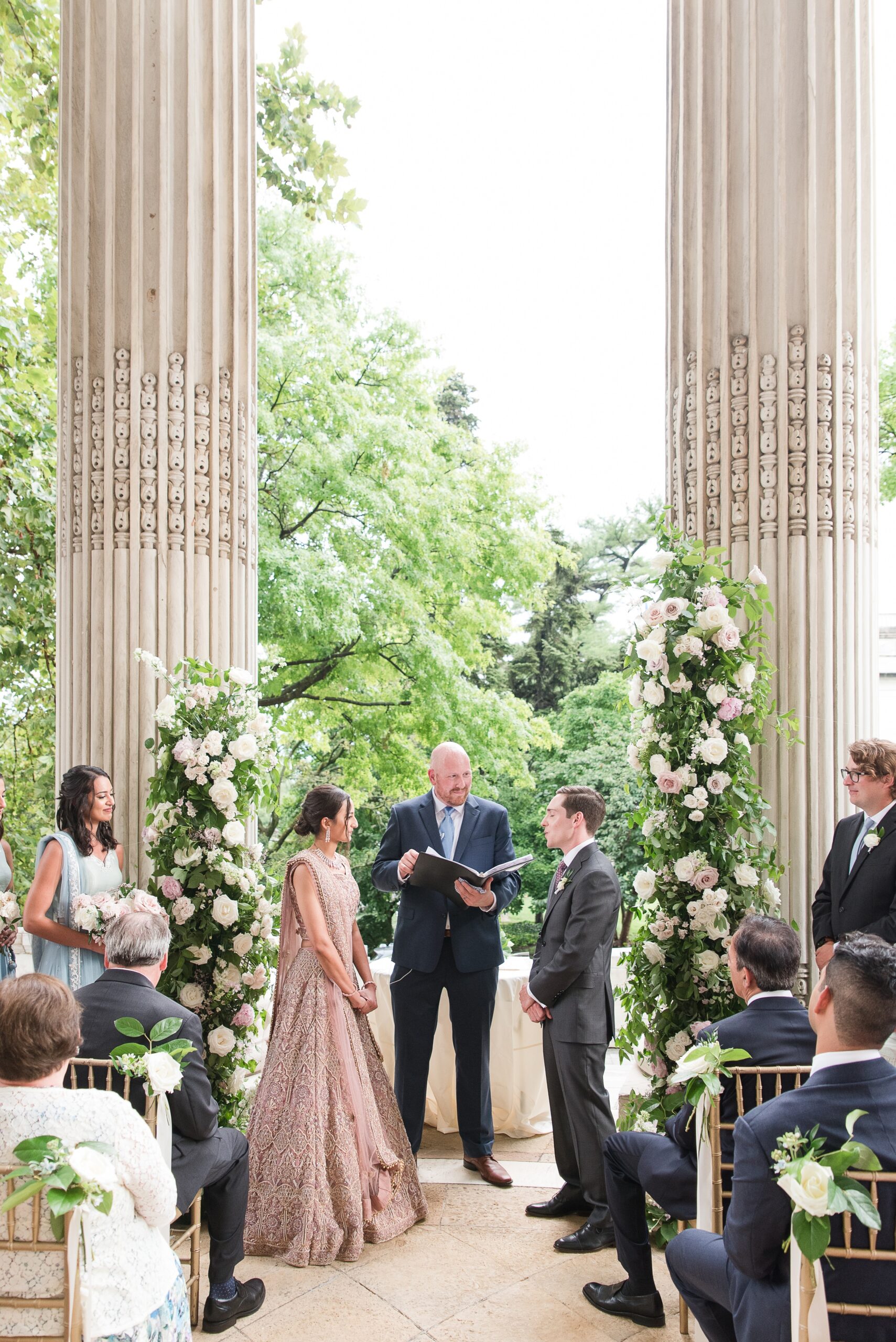 Newlyweds stand under a flower arch and ornate columns during their outdoor DAR Wedding ceremony