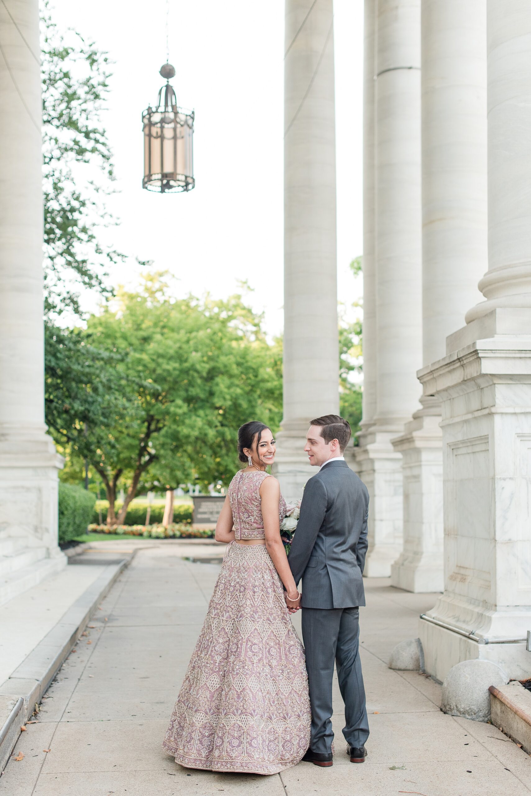 Newlyweds hold hands while smiling and walking through large ornate columns at their DAR Wedding