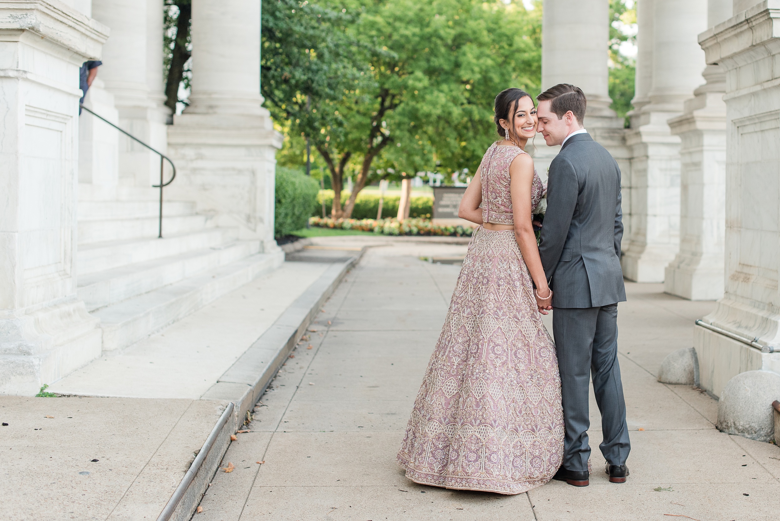 A bride smiles while her groom nuzzles her cheek and holds her hand while walking through large columns