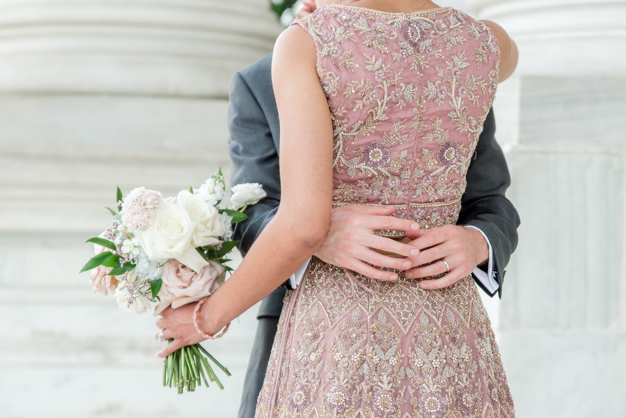 Details of a bride's ornate pink dress while she is hugged by her groom